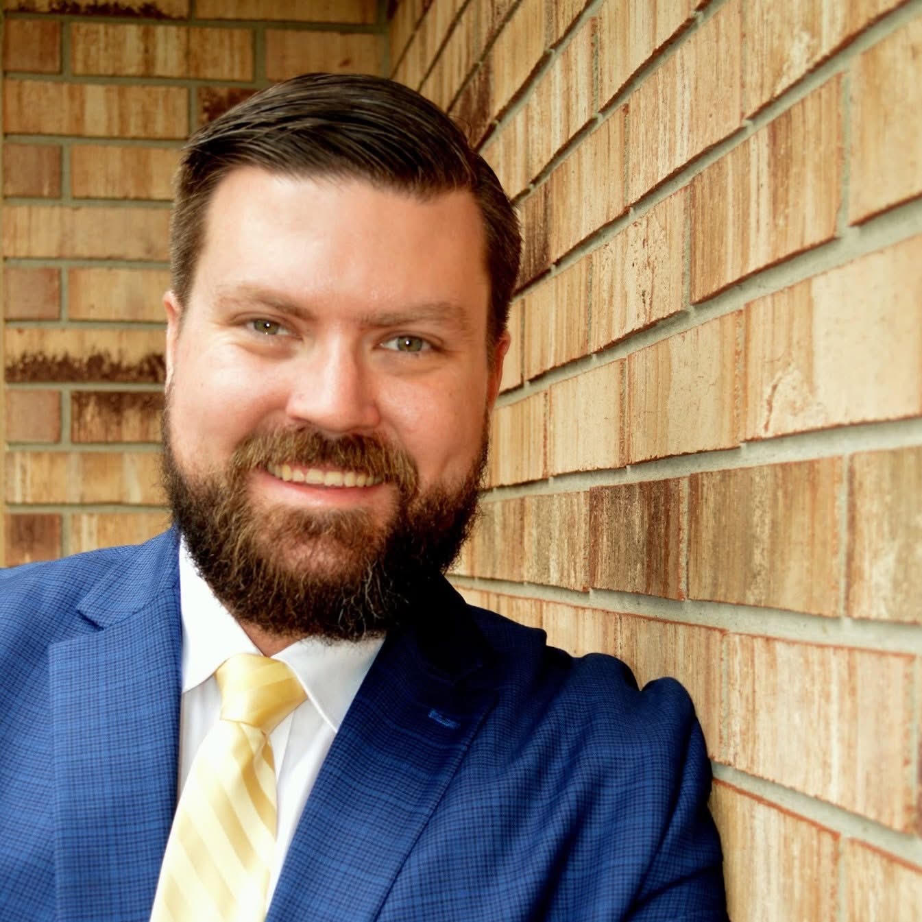 Andrew, in a blue suit jacket and gold striped tie, smiling against a brick wall